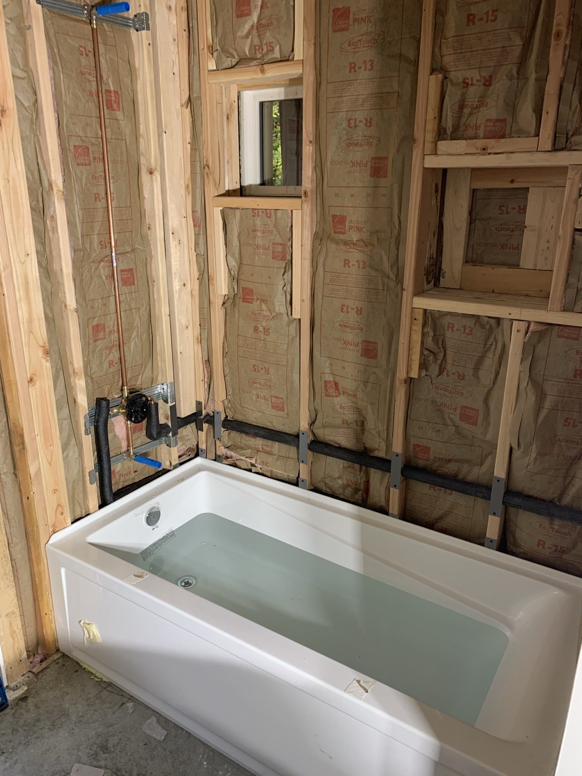 Bathtub beneath exposed shower valves and pipework in an unfinished wall