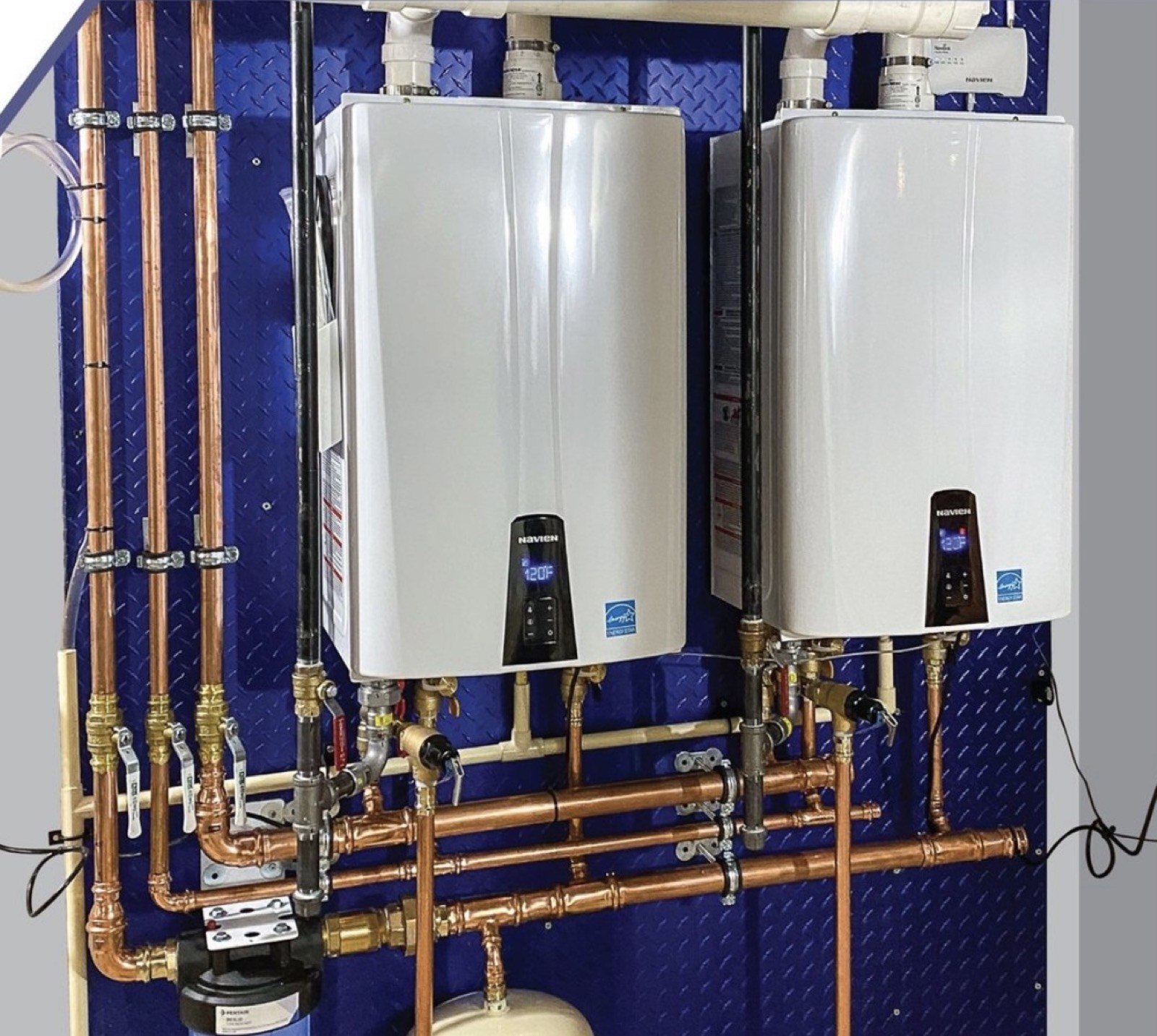 Two Navien tankless water heaters with copper pipework on a blue mounting panel